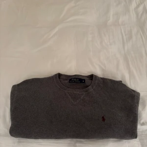 Grå stickad tröja Polo Ralph Lauren - Snygg grå stickad tröja från Polo Ralph Lauren i storlek M. Klassisk rund hals och långärmad modell med den ikoniska röda logon broderad på bröstet. Perfekt för dig som gillar stilrena och tidlösa plagg med skön passform.