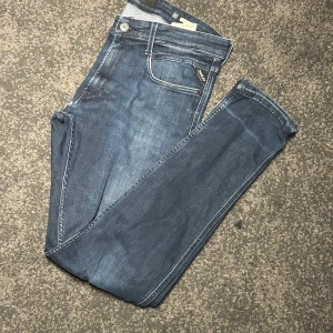 Replay Anbass mörkblå jeans herr - Prisa kan diskuteras. Snygga mörkblå Replay Anbass jeans med klassisk femficksdesign och diskreta slitningar. Slim passform och raka ben, tillverkade i mjukt jeansmaterial med stretch för extra komfort. Perfekt för dig som gillar stilrena och moderna jeans.