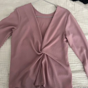 Ljusrosa omlottopp med lång ärm - Säljer en ljusrosa omlottopp med lång ärm och snygg twistad detalj framtill. Toppen har v-ringad hals och är gjord i ett mjukt, strukturerat material som känns skönt mot huden. Perfekt för dig som gillar stilrena och trendiga plagg.