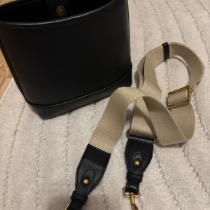 Svart axelväska från mango - Stilren svart axelväska från mango i slätt skinn med magnetlås i guld och justerbar beige axelrem med svarta skinn- och gulddetaljer. Väskan har ett rymligt fack och en innerficka för småsaker. Perfekt för dig som gillar minimalistisk design.