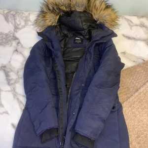 Mörkblå parkas från Everest med fuskpäls - Säljer en mörkblå parkas från Everest med stor huva och fuskpäls. Jackan har långa ärmar, dragkedja och knappar framtill samt flera fickor. Insidan är quiltad och fodrad för extra värme. Perfekt för kalla dagar och snygg stil. I storlek S