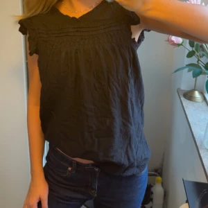 Svart blus med volangärmar - Säljer en svart blus med korta volangärmar och rundad hals. Blusen har fina rynkade detaljer upptill och är tillverkad i ett lätt, luftigt tyg som ger en skön känsla. Perfekt att styla med jeans eller kjol för en chill look. Blusen är knappt använd och där av i väldigt bra skick!!💕💕