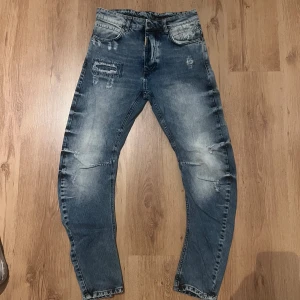 Blå jeans från Adrian Hammond med slitningar Size 30 - Säljer ett par helt sjuka blå jeans från Diamond med coola slitningar och broderade detaljer på både fram- och baksida. Jeansen har normal passform, fem fickor och klassisk knappgylf. Färgen är tvättad blå med ljusare partier och materialet är robust denim. Midja 38cm längd 102cm (68) 