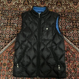 Svart quiltad dunväst från Kappahl - Quiltad svart dunväst från Kappahl med blått foder, två dragkedjefickor och hög krage. Västen har ett snyggt diamantmönster och en liten bröstficka med knapp. Perfekt för lager-på-lager och chill stil.