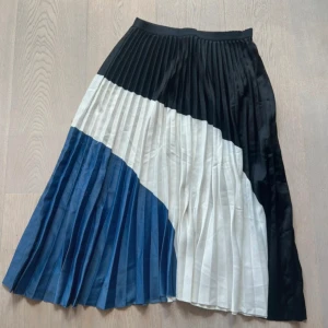  Långkjol H&M - Snygg plisserad långkjol från H&M med cool färgblockad design i svart, vitt och blått. Kjolen har bred resår i midjan och är tillverkad i ett lätt, glansigt material som ger ett stilrent intryck. Perfekt för dig som gillar att sticka ut med din outfit.