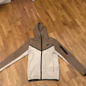 Beige och vit Nike hoodie med dragkedja - Nike hoodie i beige och vitt med svart logga på bröstet. Tröjan har hel dragkedja framtill, huva och en praktisk ficka med dragkedja på ena ärmen. Materialet är mjukt och känns som polyester. Snygg kontrast med svarta detaljer och sportig look.
