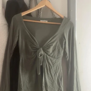 Mörkgrön långärmad topp Design by Si - Snygg mörkgrön långärmad topp från Design by Si i mjuk bomull. Modellen har djup v-ringning och en cool knyt-detalj framtill som ger en feminin touch. Passformen är slim och toppen är perfekt att styla med jeans eller kjol för en trendig look.