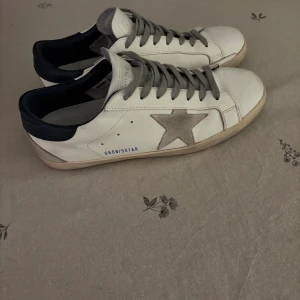 Golden Goose vita sneakers - Golden Goose sneakers i vitt skinn med grå mockadetaljer och ikonisk stjärna på sidan. Grå snörning, mörkblå häl och platt sula. Snygg, avslappnad look med vintage-vibe. Kommer med kartong, qr kod och dustbag.