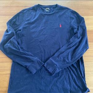Marinblå långärmad tröja Polo Ralph Lauren - Snygg marinblå långärmad tröja från Polo Ralph Lauren med klassisk röd broderad logga på bröstet. Tröjan är i mjuk bomull och har rund hals samt ribbade muddar vid ärmslut. Perfekt för dig som gillar stilrena och tidlösa plagg.