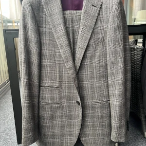 Grå rutig kostym från Mabro - Snygg grå kostym från Mabro, handgjord i Toscana, Italien. Kavajen har klassisk passform med två framfickor och knappar, och byxorna har pressveck. Rutigt mönster i olika grå nyanser ger en stilren vibe. Perfekt för dig som gillar italiensk design och vill sticka ut med en tidlös look. Använd bara 2-3 gånger.