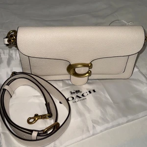 Vit axelväska från coach tabby  - Snygg axelväska från Coach i ljusbeige skinn med justerbar rem och guldfärgade metalldetaljer. Väskan har ett stort C-spänne framtill, innerfack med dragkedja och stilren design. Perfekt för dig som gillar klassiska och trendiga accessoarer.