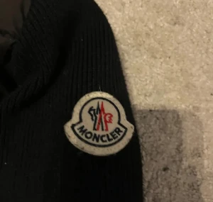 Svart Moncler pufferjacka med stickade ärmar - Svart pufferjacka från Moncler med stickade ärmar och hög krage. Jackan har dunfylld, quiltad front och dragkedja framtill. Klassisk Moncler-logga på ärmen. Materialmix av ull och dun ger både värme och stil.