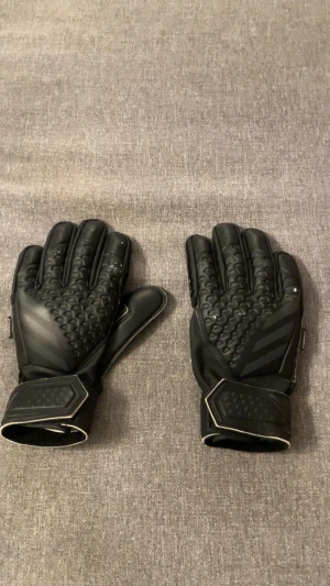 Svarta målvaktshandskar från Adidas - Säljer ett par svarta målvaktshandskar från Adidas med coolt mönstrad ovansida och släta insidor. Handskarna har justerbara muddar med kardborre och diskreta Adidas-loggor. Materialet är syntet och designen är sportig med extra grepp på ovansidan. Använd 1-2 träningar men är i väldigt fint skick!
