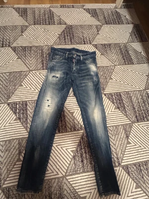 Blå Dsquared2 jeans med slitningar - Snygga blå jeans från Dsquared2 med coola slitningar och tvättade detaljer. Klassisk femficksmodell med knappstängning och Dsquared2-logga på röd etikett vid gylfen. Jeansen har en rak passform och är tillverkade i jeansmaterial.