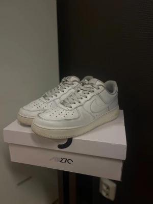 Nike Air Force 1 - Fräscha vita Nike Air Force 1 sneakers med klassisk låg silhuett och rund tå. Skorna har snörning, perforerade detaljer på tån och är tillverkade i skinn med vit sula. Perfekt för dig som gillar stilrena och ikoniska sneakers.