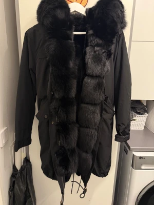 Svart parkasjacka med fuskpäls - Säljer en svart parkasjacka med stor fluffig fuskpäls längs huva och insida. Jackan har dragsko i midjan och nederkant, samt praktiska fickor och långa ärmar. Perfekt för kalla dagar när du vill vara både varm och snygg.