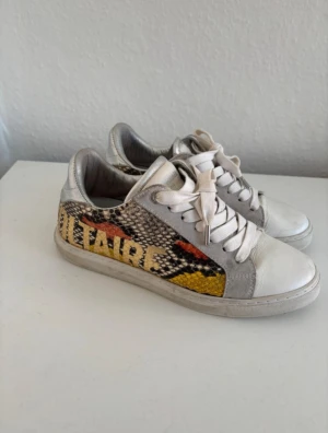 Zadig & Voltaire sneakers med mönster - Snygga sneakers från Zadig & Voltaire med coolt ormmönster i svart, gul och röd på sidorna. Skorna har vit och silverfärgad ovandel i skinn och mocka, samt vita snören och platt sula. Perfekt för dig som gillar statement-detaljer och streetstyle.