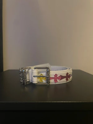 Vitt skärp med färgglada kors - Säljer ett vitt bälte med silverfärgat spänne och detaljerade kors i gult, rosa och rött längs hela bältet. Skärpet är gjort i skinn och har en unik, edgy vibe med sina färgglada kors och exklusiva känsla.