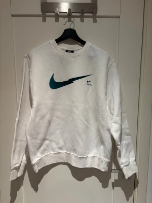 Vit Nike sweatshirt med blå logga - En clean vit sweatshirt från Nike i standard fit med stor blå blixtformad swoosh-logga och trycket 20/21 på bröstet. Tröjan är använd en gång, i stort sätt helt ny utan prislapp.