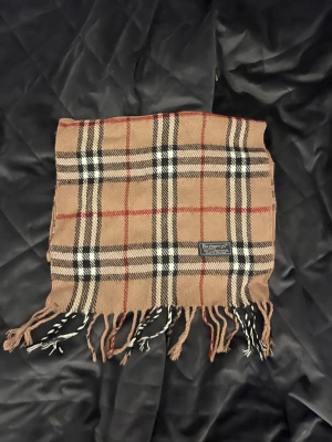 Rutig beige halsduk från Burberry - Snygg klassisk halsduk från Burberry i beige med rutigt mönster i svart, vitt och rött. Gjord av 100% lammull och har fransar i ändarna. Perfekt för att ge din outfit en lyxig touch och hålla dig varm under kyliga dagar.