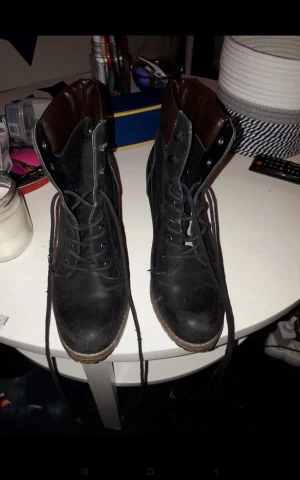 Svarta stövletter med snörning och klack - Säljer ett par svarta stövletter med brunt foder och snygg snörning framtill. Skorna har rund tå, platt sula framtill och en hög klack i trälook. Materialet är troligtvis syntet eller skinnimitation. Perfekta för dig som vill ha en edgy men stilren look.