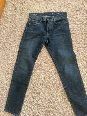 Mörkblå skinny jeans från G-Star - Snygga mörkblå jeans från G-Star med klassisk femficksdesign och skinny passform. Jeansen har en stilren look med svarta detaljer på bakfickan och ett svart läderpatch med G-Star-logga i midjan. Perfekt för dig som gillar en smal siluett och street-inspirerad stil.