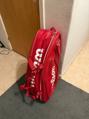 Wilson röd tennisväska med dragkedja - Stor och sportig tennisväska från Wilson i rött med vita logotyper. Väskan har flera fack med dragkedjor, justerbara axelremmar och extra handtag. Perfekt för att få plats med racket, skor och annan utrustning. Snygg design med TOUR-detalj på sidan.