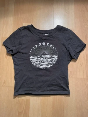 Svart t-shirt med månfaser från H&M - Cool svart t-shirt från H&M med ett stort tryck på bröstet som visar månfaser och vågor i vitt. T-shirten är croppad och har rund hals samt korta ärmar.