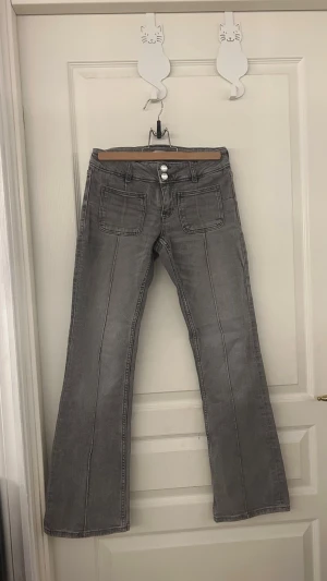 Grå bootcut jeans från Gina Tricot - Snygga grå jeans från Gina Tricot i bootcut-modell med hög midja. Jeansen har två stora fickor fram, dubbla knappar i midjan och klassiska bakfickor. Materialet är mjukt denim och färgen är mörkgrå med lätt tvättad look.