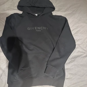 Svart Givenchy hoodie med huva - Svart hoodie från Givenchy med stor logga på bröstet. Tröjan har huva, ribbade muddar och en klassisk känguruficka framtill. Materialet är mjukt och känns skönt mot huden, perfekt för dig som gillar stilrena designer och exklusiva märken.