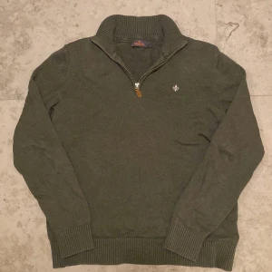 Mörkgrön stickad tröja Morris half zip - Snygg stickad tröja från Morris med half zip och ribbad krage i färgen Oliv. Tröjan har långärmad passform och en broderad Morris logga på bröstet. Perfekt för dig som gillar klassisk stil med en modern touch.