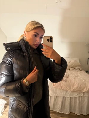 Svart lång pufferjacka BOSS - Säljer en svart, lång pufferjacka märke BOSS med hög krage och huva. Jackan har glansig yta, dragkedja hela vägen och breda, vadderade sektioner. Sitter snyggt med en loose passform och är perfekt för kalla dagar. Enkel och stilren design som funkar till allt. Köptes för 6000kr aldrig använd 