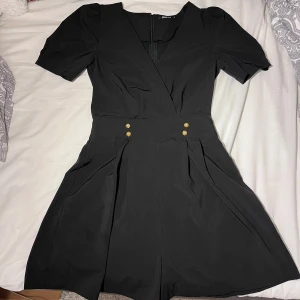 Svart shortsdress - En shortsdress från Gina Tricot med guldfärgade knappar framtill. Modellen har omlott-look upptill och dragkedja bak. Perfekta för dig som gillar en klassisk och chic vibe.