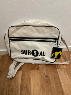 Vit axelväska från Daniel ray - Vit axelväska från Daniel ray med svart tryck och svarta detaljer. Väskan har justerbar axelrem, dragkedja framtill och flera fack inuti. Tillverkad i syntetmaterial med en clean och sportig look. Perfekt för att bära med sig allt man behöver till skolan eller stan. (Ordinarie pris: 1500kr)