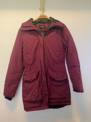 Vinröd parkasjacka från Everest - Parkasjacka i vinröd/lila färg från Everest med fuskpäls på huvan och stora fickor framtill. Jackan har dragkedja och knappar, lång passform och är helfodrad. Underbar jacka. Otroligt varm. Hade den fortsatt känts hemma på mig, hade jag behållt den. Missfärgade vita fläckar främst på ena ärmen och framtill vid fickorna, troligtvis ifrån användning av fel tvättmedel samt ett litet hål i en av framfickorna men utöver det - är den i så fint skick! 