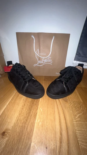 Christian Louboutin svarta sneakers mocka - Säljer ett par svarta sneakers från Christian Louboutin i lyxig mocka med klassisk röd sula och diskret logga. Skorna har rund tå, svarta snören och detaljer i svart. Insidan är i beige skinn för extra komfort. Perfekta för dig som vill sticka ut med stil.