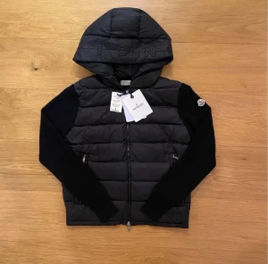 Svart pufferjacka från Moncler med huva - Snygg svart pufferjacka från Moncler med vadderad kropp och stickade ärmar. Jackan har huva med broderad logga, dragkedja framtill och Moncler-märke på ärmen. Perfekt för kyliga dagar och ger en clean, exklusiv look.