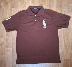 Brun pikétröja Polo Ralph Lauren L - Brun pikétröja från Polo Ralph Lauren i storlek L. Tröjan har klassisk krage, korta ärmar och två knappar framtill. Stor broderad polospelare på bröstet och ett stort broderi med polospelare på ryggen. Snygg kontrastdetalj på ena ärmen. Materialet är mjuk bomull. OBS JAG HAR LIMMAT UPP TRÖJAN MED BOMULLS LIM 8,5 CM!