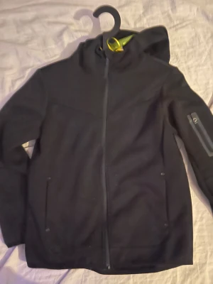 Svart fleecejacka från Primark - Svart fleecejacka från Primark med huva och hel dragkedja framtill. Jackan har två sidofickor och en extra ficka med dragkedja på ärmen. Perfekt för kyliga dagar och enkel att matcha med allt.