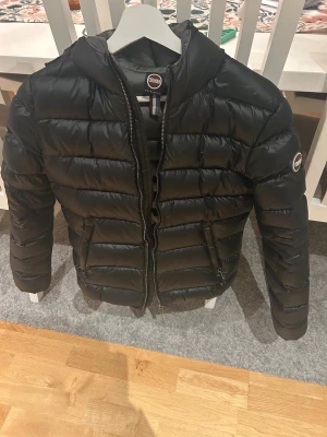 Svart dunjacka från Colmar - Snygg svart dunjacka från Colmar med quiltad design och dragkedja framtill. Jackan har en klassisk pufferstil, lång ärm och två fickor med dragkedja. Perfekt för kyliga dagar och ger en clean look till din outfit. Har tre små håll som är täckta och syns inte vid användning (närmare bilder 2,3) ena sitter bak vid armen som inte syns överhuvudtaget och andra fram som inte heller syns så mycket då den svarta färgen sjunker in i jackan. Storlek 14y skulle säga att det motsvarar ca xs,s. Nypris 5000