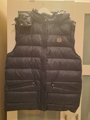 Mörkblå dunväst från Bvaldi, storlek L - Mörkblå dunväst från Bvaldi med huva och två fickor med dragkedja. Nypris: 1799kr. Mycket fint skick.