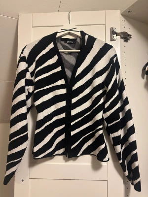 Zebramönstrad kofta från Gina Tricot - Snygg zebramönstrad kofta från Gina Tricot i svart och vitt. Koftan har lång ärm, v-ringad hals och knappar framtill. Perfekt för dig som vill sticka ut med ett djuriskt mönster och cool stil. Materialet är mjukt och känns skönt mot huden.