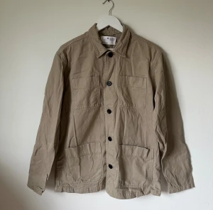 Beige overshirt från Selected Homme - Stilren beige overshirt från Selected Homme i 100% bomull. Jackan har klassisk krage, fyra stora fickor framtill och knäppning med svarta knappar. Perfekt lager-på-lager-plagg med avslappnad passform och lång ärm. 4Ä5