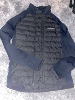 Svart quiltad jacka Peak Performance S - Svart quiltad jacka från Peak Performance i storlek S. Jackan har släta ärmar och sidor i stretchigt material, quiltad front och rygg, samt dragkedjor på fickorna. Perfekt för dig som gillar sportig och clean stil.