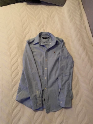 Blå skjorta från Ralph Lauren XS - Klassisk ljusblå skjorta från Ralph Lauren i custom fit. Skjortan har långa ärmar, knappar framtill och den ikoniska broderade polospelaren på bröstet. Tillverkad i bomull och passar perfekt för en clean och stilren look.