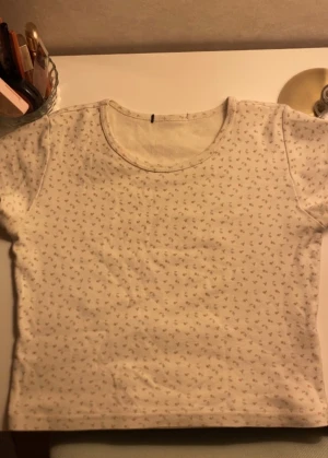 Blommig t-shirt - Croppad t-shirt i färgen creme med små blommiga mönster i pastellfärger. Tröjan ger ”90’s baby tee” vibes fast lite mer croppad