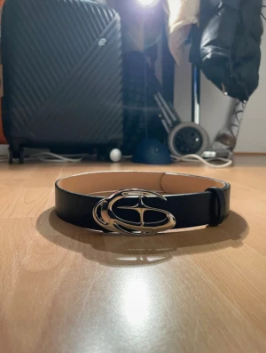 Svart skärp med silverfärgat spänne - Svart bälte i fejk skinn med ett stort, unikt silverfärgat spänne framtill. Passar till jeans och andra byxor eller kjolar. Insidan är ljusbeige och bältet är brett för att sticka ut som accessoar.