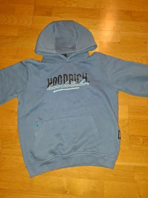 Blå hoodie från Hoodrich med huva - Snygg blå hoodie från Hoodrich med stor logga på bröstet och coolt tryck i svart och vitt. Tröjan har huva, lång ärm och en praktisk ficka med dragkedja framtill. Perfekt för chill dagar och streetwear-stil.