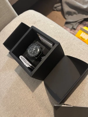 Omega swatch snoppy  - Säljer en stilren svart Omega swatch Speedmaster med rund boett och tachymeter-skala. Klockan har svart urtavla med vita detaljer och tre subdials. Armbandet är svart med Omega-logga. Perfekt för dig som gillar klassisk design och ikoniska märken. Öppen för byten med. 