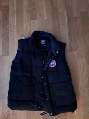 Svart dunväst från Canada Goose - Svart dunväst från Canada Goose med klassisk logopatch på bröstet. Västen har två stora fickor framtill, knappar och dragkedja. En väldigt gammal model på västen. Knapparna färg har försvunnit lite men pris kan diskuteras 